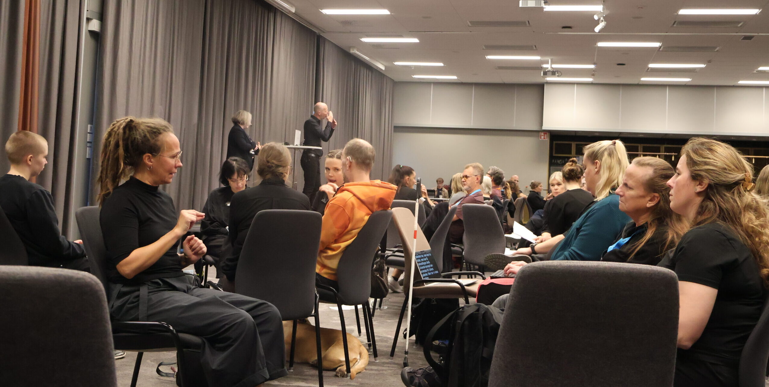 Deltagare sitter i mindre grupper och samtalar på teckenspråk i en konferenslokal. I förgrunden syns en kvinna som tecknar och en liggande hund.