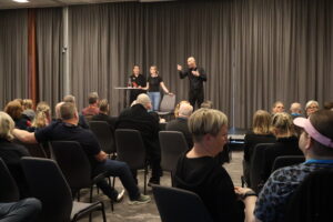 Medlemmarna sitter i rader av stolar i konferenslokalen. Längst fram står en tolk.  Bredvid honom står Sophie Toomväli Peterson och föreläser.