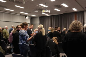 Syntolkning: Medlemmar och tolkar står upp i konferenslokalen. I lokalen finns rader av grå stolar, mörka gardiner och takbelysning.